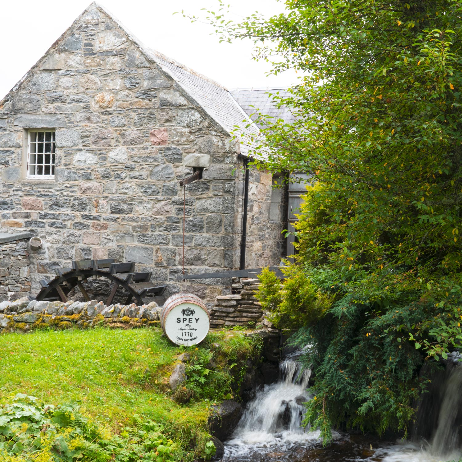 Historische steinerne Wassermühle mit Wasserrad und grünem Garten, nahe einem kleinen Wasserfall und einem Fass mit "SPEY" Aufschrift.