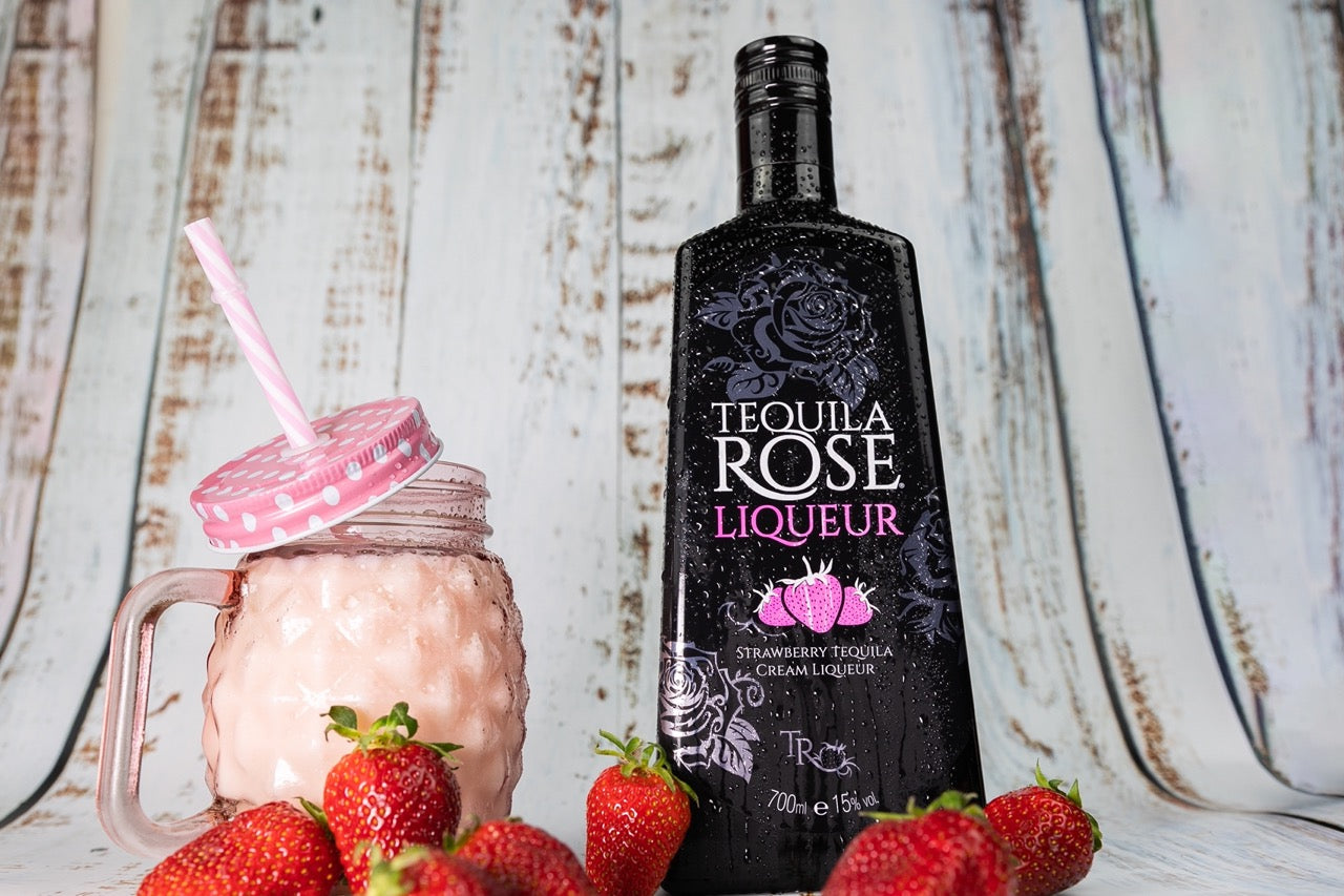 Eine Tequila Rose Liqueur-Flasche neben einem pinken Getränk in einem Glas mit Erdbeeren auf einem rustikalen Hintergrund.